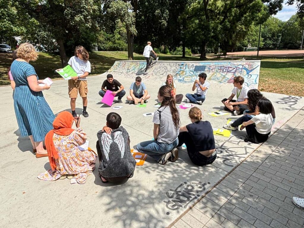 Teilnehmende des Workshops zu Jugendsprache sitzen an einer Skateanlage im Kreis und arbeiten gemeinsam mit bunten Plakaten.